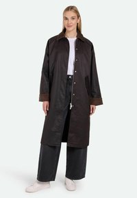 Lang mørkebrun frakke med et kordfløjlskragen, trykknapper og sidelommer, båret over en hvid top og brede sorte jeans.