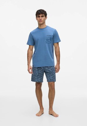 Giovane uomo in piedi a piedi nudi che indossa una maglietta blu a maniche corte e pantaloncini blu a fantasia su sfondo bianco neutro.