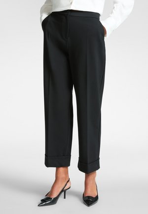 Schwarze Hose mit hohem Bund, Falten und umgeschlagenen Säumen, aus glattem Stoff. Kombiniert mit schwarzen spitzen Slingbacks.