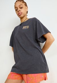 Mørkegrå oversized T-skjorte med et vintage-logo, kombinert med lyse rosa rutete shorts med et subtilt mønster.