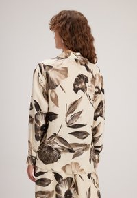 Blouse à manches longues en tissu léger, avec un motif floral dans des nuances de marron sur un fond crème ; boutons discrets aux poignets.