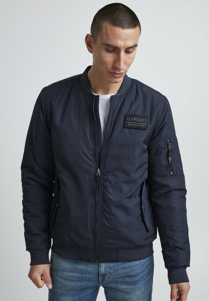 Chaqueta bomber de color azul marino fabricada con una tela suave, con cierre de cremallera en el frente, puños acanalados y una etiqueta de parche en el frente. Bolsillo en la manga izquierda.