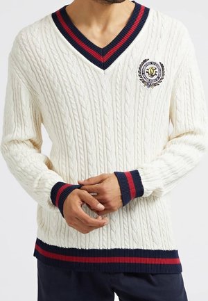 Mand iført hvid kabelstrikket V-hals sweater med marineblå og røde striber på krave, manchetter og kant, med en broderet våbenskjold på brystet.