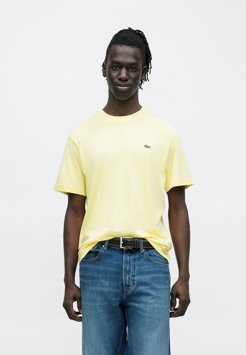 T-shirt di cotone giallo con scollo rotondo e maniche corte, con un piccolo logo verde sul petto. Abbinata a jeans blu e una cintura nera.