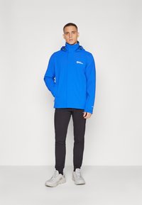 Veste imperméable bleu avec une fermeture éclair à l'avant, une capuche ajustable et un détail de logo. Associée à un pantalon noir ajusté et des chaussures de sport grises.