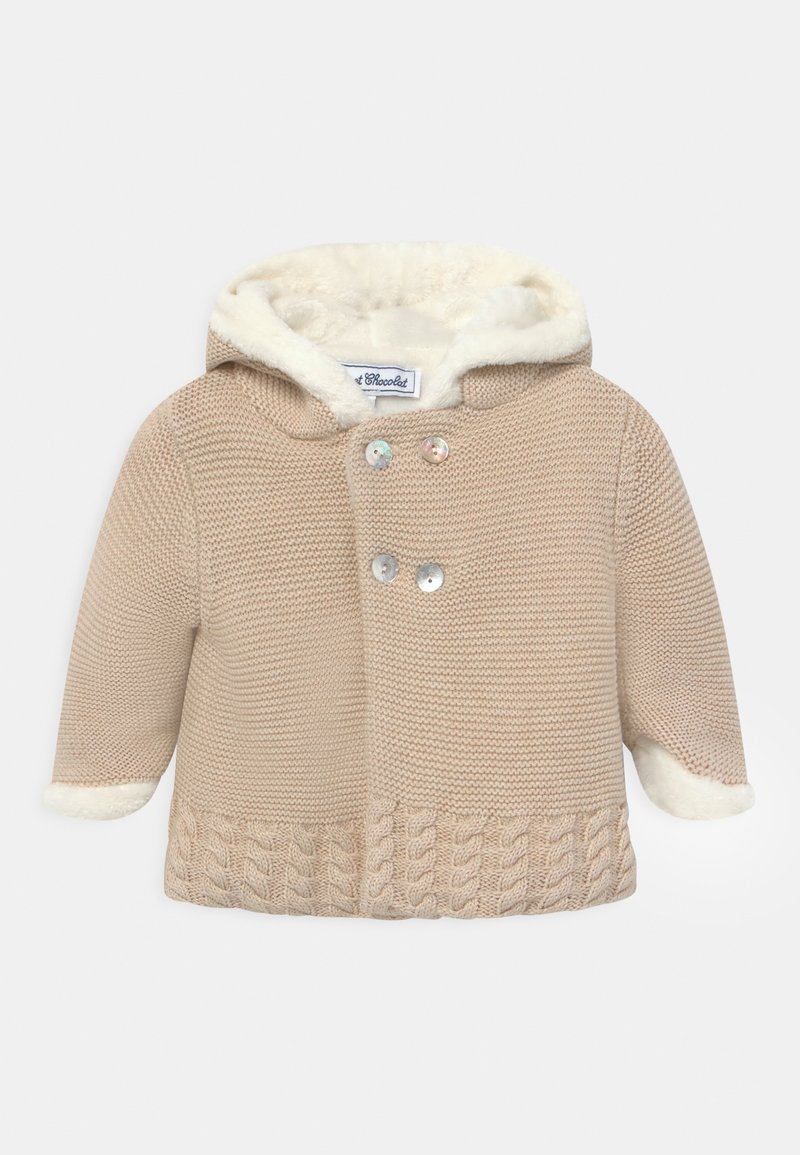 Beige stickad kappa med en foder av päls i huvan. Har ett flätat mönster vid nederkanten och två iriserande knappar för stängning.