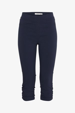 Femeie purtând pantaloni conici bleumarin și un top alb cu o brățară bej la încheietură, mâna sprijinită pe șold.