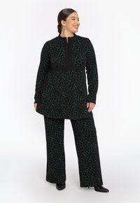 Lachende vrouw in een zwart en groen geblokte lange tuniek over bijpassende wijde broek en zwarte enkellaarsjes, staand tegen een witte achtergrond.