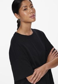 Svart bomullströja med korta ärmar, rund halsringning och avslappnad passform. Modellen har håret i en låg hästsvans och armarna i kors.