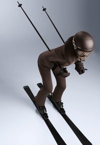 Hnedý lyžiarsky oblek s kožušinovým golierom, čierne lyžiarske vybavenie a lyžiarske palice. Lyžiar sa nakláňa dopredu na čiernych lyžiach, s aerodynamickou prilbou a okuliarmi.