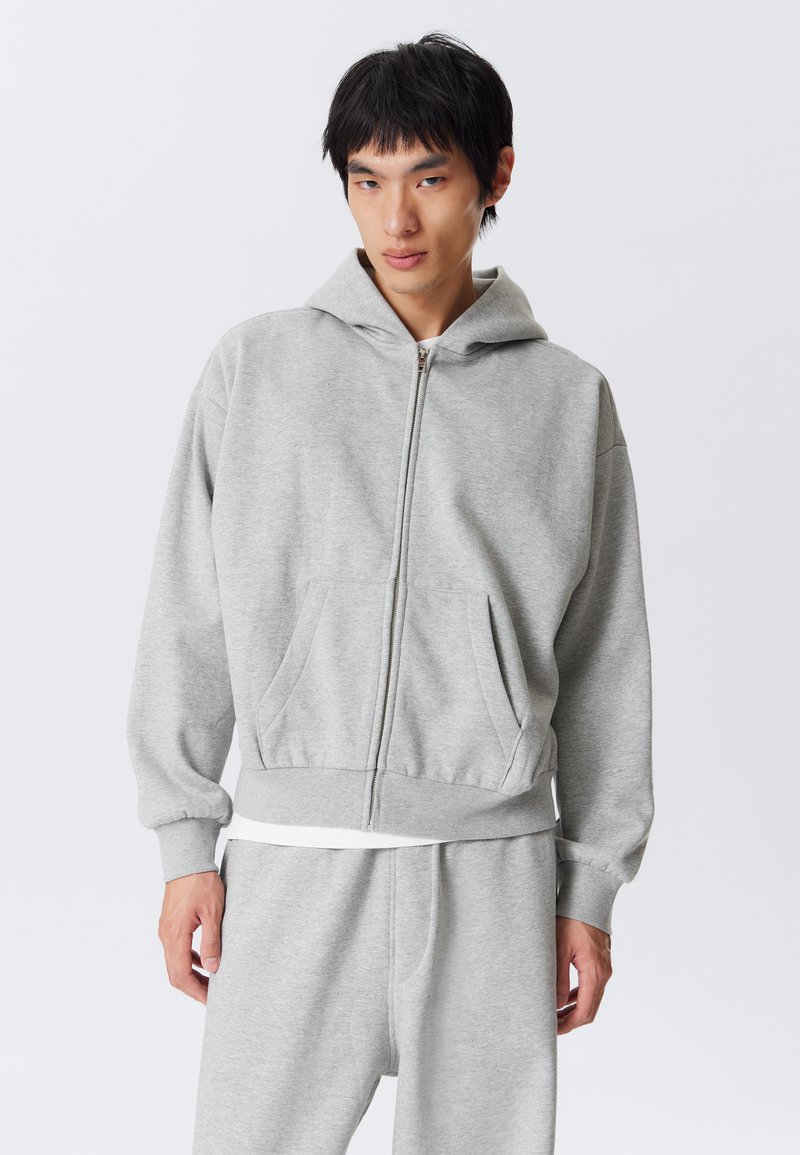 Grå zip-up hoodie med en frontlomme, ribbede ærmer og kant. Lavet af blødt stof med en afslappet pasform. Kombineret med matchende grå sweatpants.