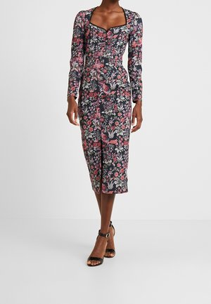 Kvinde iført en tætsiddende midi-kjole med lyserøde og hvide blomster- og fugleprint, lange ærmer, firkantet halsudskæring og sorte ankelstrophæle.