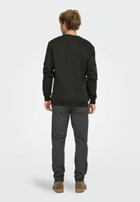 Homme debout de dos portant un sweat-shirt noir, un pantalon gris foncé et des bottes en daim marron sur un fond clair uni.
