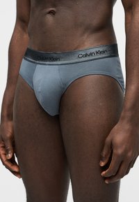 Férfitörzs szürke Calvin Klein alsónadrágban, márkázott derékrésszel, egyszínű, világos háttér előtt állva.