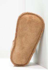 Pantufla de ante beige con punta redondeada y bordes cosidos, que presenta una textura suave y una suela plana, situada sobre un fondo blanco.