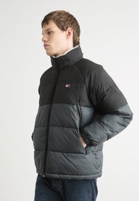 Giacca a vento nera e grigia con collo alto, chiusura con zip e tasche laterali. Presenta ampie righe orizzontali e dettaglio del logo sul petto.