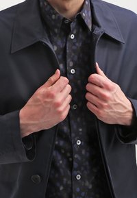 Muž upravuje límeček tmavě námořnického kabátu, který má na sobě černou ko�šili s jemným abstraktním vzorem modré a fialové barvy.