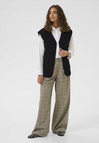 Crni vuneni prsluk s ovratnikom i džepovima, uparen s bež kariranim hlačama širokih nogavica i crnim cipelama bez vezivanja s dekorativnim čavlima.