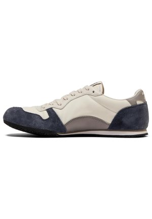 Sneaker featuring a beige leather upper with navy suede accents, a curved design, lace-up closure, and a black rubber sole.