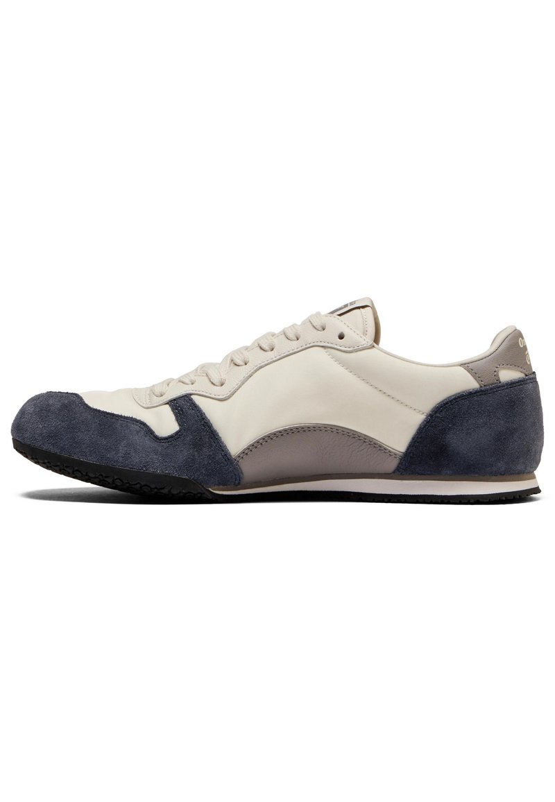 Sneaker z beżową skórzaną cholewką, z granatowymi zamszowymi akcentami, zakrzywionym kształtem, sznurowanym zapięciem i czarną gumową podeszwą.