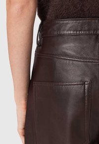 Brūni ādas bikses ar gludu tekstūru, kurām ir plata jostasvieta, jostas cilpas un viena aizmugures kabata. Tīri šuvju akcenti.