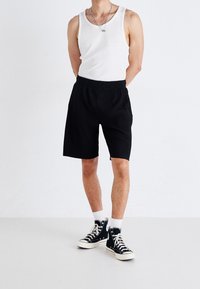 Shorts negros de canalé con cinturilla elástica, combinados con una camiseta sin mangas blanca y zapatillas altas negras. Fondo de estudio.