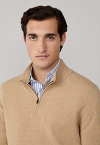 Giovane uomo con capelli scuri che indossa un maglione beige a collo zip parziale sopra una camicia a righe blu e bianche, con uno sguardo neutro rivolto dritto davanti a sé.