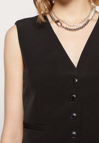 Blazer noir sans manches avec un décolleté en V profond, cinq boutons noirs, poches latérales et une texture de tissu lisse. Accentué par des colliers superposés.