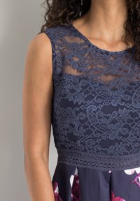 Modré krajkové šaty s květinovým vzorem a průhledným výstřihem. Hladká tkanina se přiléhavým trupem a rozšířenou sukní. Podrobná krajková svrchní vrstva.