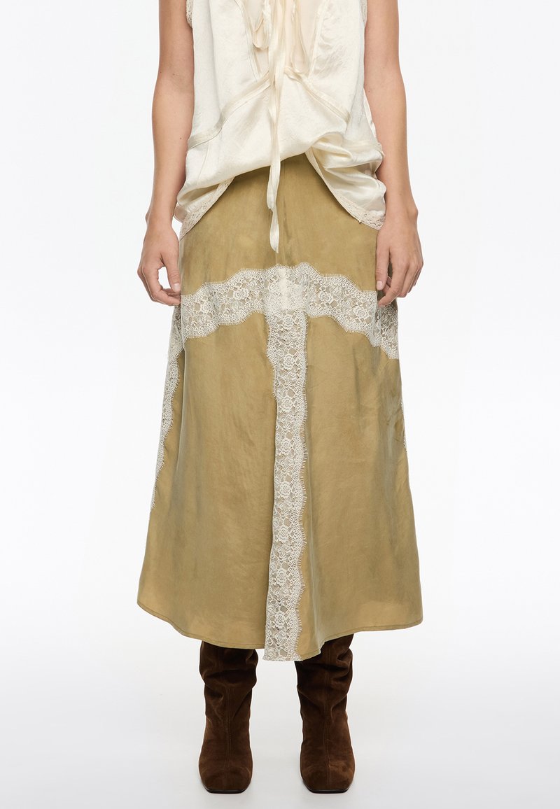Falda midi verde oliva con borde de encaje, que presenta un diseño fluido y una textura suave. Combinada con botas de ante marrón hasta la rodilla.
