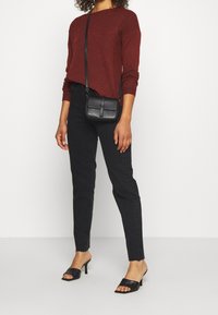 Roter Strickpullover, schwarze Slim-Fit-Jeans, schwarze Handtasche mit Trageriemen und schwarze Sandalen mit offenem Zehenbereich. Glatte Textur, figurbetontes Design und neutrale Farben.