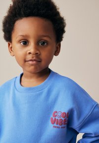 Sweatshirt en coton bleu avec un col côtelé, arborant la mention "COOL VIBES" et un graphique de visage souriant en rouge. Texture douce, design décontracté.