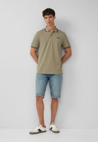 Oliwkowa koszulka polo z czarno-białym prążkowanym kołnierzykiem, zestawiona z jasnoniebieskimi szortami dżinsowymi. Białe sneakersy z czarnymi akcentami.