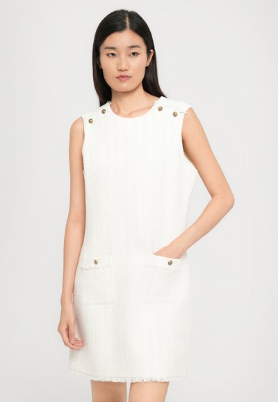 Biała bezrękawnikowa sukienka z teksturowanej tkaniny, z dwoma przednimi kieszeniami z złotymi guzikami oraz frędzlami na hemie.