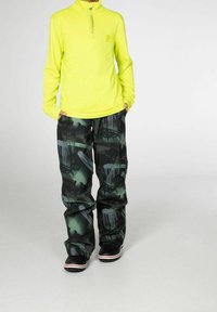 Top amarillo neón de manga larga con cuello con cremallera, combinado con pantalones holgados de patrón verde y negro con diseños abstractos. Zapatos negros.
