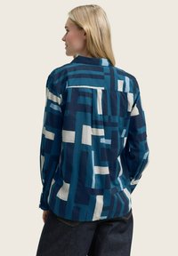 Camicia a fantasia blu con disegni geometrici in diverse tonalità di blu e bianco. Il tessuto sembra leggero e presenta un colletto con bottoni.