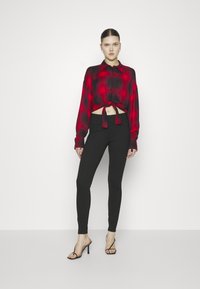 Chemise crop rouge et noire à carreaux avec un nœud à l'avant, portée avec des leggings noirs taille haute et des talons aiguilles noirs à lanières. Textures lisses.