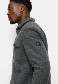 Gray wool-blend jacket with a collared neckline, featuring a button-up front, single chest pocket, and a black logo patch on the sleeve.