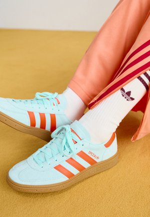 Jasnoniebieskie sneakersy Adidas Spezial z pomarańczowymi paskami, noszone z białymi skarpetkami Adidas i brzoskwiniowymi spodniami dresowymi na żółtej podłodze.