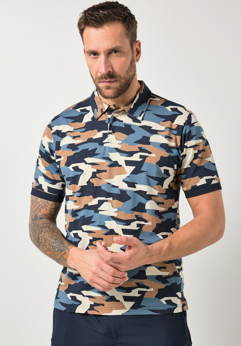 Mann mit tätowiertem Arm, der ein kurzärmeliges Poloshirt in Blau, Beige, Schwarz und Weiß im Tarnmuster vor neutralem Hintergrund trägt.