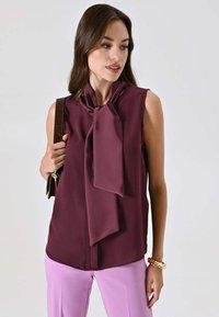 Blusa bordeaux senza maniche con dettaglio a fiocco sul collo, texture liscia e vestibilità ampia, abbinata a pantaloni rosa chiaro.