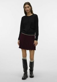 Cardigan noir en tricot avec un col rond et une fermeture à boutons ; associé à une jupe plissée bordeaux et des bottes noires jusqu'aux genoux.