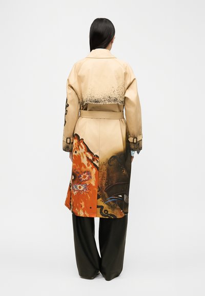 Γυναίκα με μακριά σκούρα μαλλιά φοράει μια μπεζ καμπαρντίνα με πορτοκαλί και μαύρα αφηρημένα σχέδια στην πλάτη, συνδυασμένη με μαύρο παντελόνι φαρδύ στο πόδι.