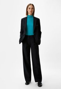 Czarna, dopasowana marynarka i spodnie z zestawione z turkusową bluzką z wysokim kołnierzem. Model stoi, ręce w kieszeniach, w czarnych butach na jednolitym tle.