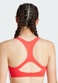 Sports bra u svijetlo crvenoj boji s dizajnom racerback, s izrezanim detaljima i bijelim trakama na stranama. Glatka tekstura tkanine.