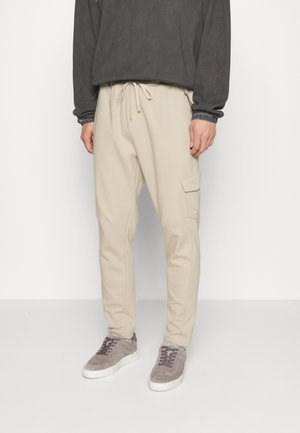 Mężczyzna w beżowych spodniach cargo z troczkiem w pasie, szarym długim rękawie i szarych zamszowych butach sportowych na białym tle.