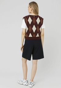 Chemise blanche à manches courtes sous un gilet en tricot argyle bordeaux et beige, associés à un short rayé noir et des baskets blanches.