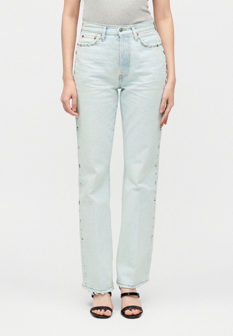 Jean droit taille haute bleu clair pour femme avec des clous métalliques le long des coutures latérales, porté avec des sandales noires à bouts ouverts et un top côtelé gris clair.