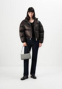 Giacca puffer marrone con design trapuntato, chiusura con zip, maglietta nera, jeans scuri e una borsa a tracolla con stampa e tracolla sottile, su sfondo bianco.