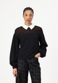 Schwarzer Sweatshirt mit einem durchsichtigen oberen Abschnitt in Schwarz und einem weißen Kragen, mit langen Puffärmeln und einer lockeren Passform. Kombiniert mit gemusterten Hosen.